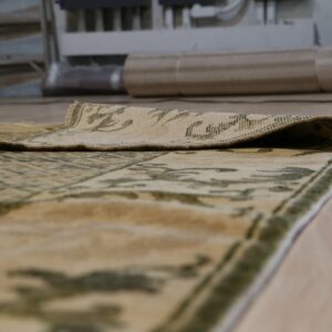 The folded edge of a beige and green patterned low-pile rug rests on light wood flooring.