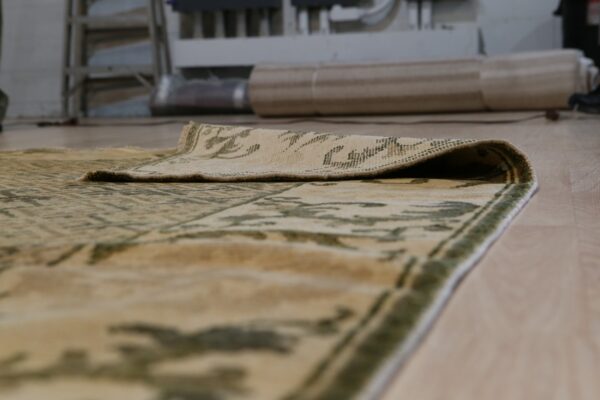The folded edge of a beige and green patterned low-pile rug rests on light wood flooring.