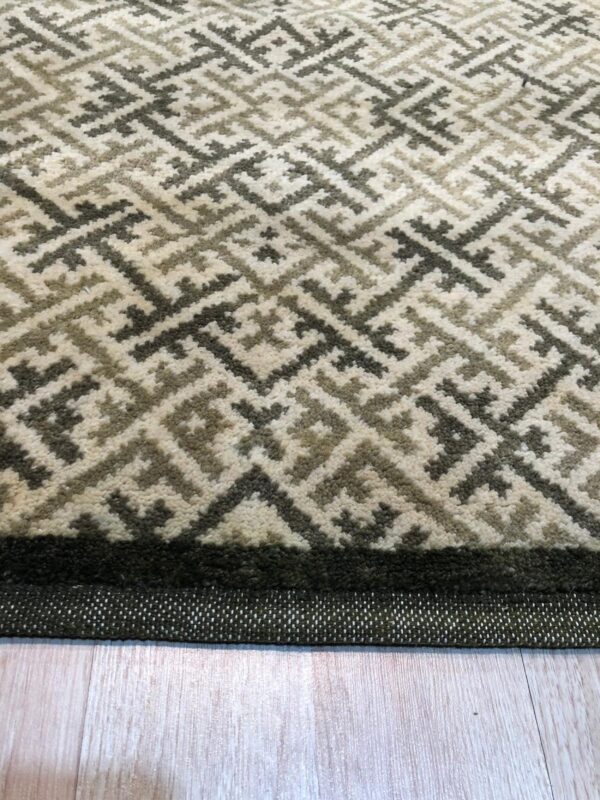 A medium pile rug with a geometric pattern in cream, gray, and tan rests on light wood planks.