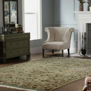 Traditional sage green area rug with dark scrolling patterns defines a living room space with a cream wingback chair.
