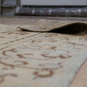 The edge of a low-pile cream and brown patterned rug is curled back over light wood flooring.
