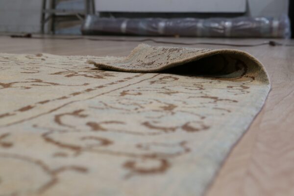 The edge of a low-pile cream and brown patterned rug is curled back over light wood flooring.