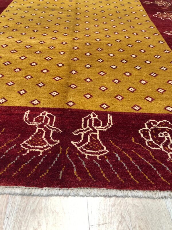 Gold rug with small geometric motifs and a maroon border showing stylized figures on a wood floor.