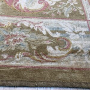 Close-up of a medium-pile rug border in khaki with pink and cream ornate floral scrollwork on light wood flooring.