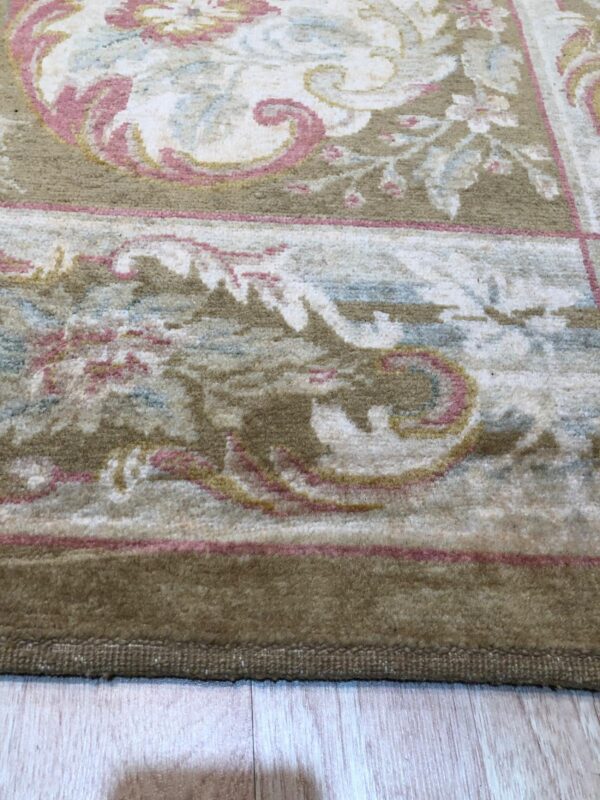 Close-up of a medium-pile rug border in khaki with pink and cream ornate floral scrollwork on light wood flooring.