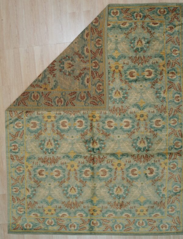 Medium pile teal rug with rust and gold traditional patterns, corner folded back on wood floor.