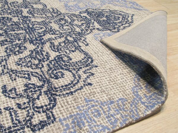 Blue ornamental rug with cream and gray accents, curled back to show the woven reverse on wood planks.