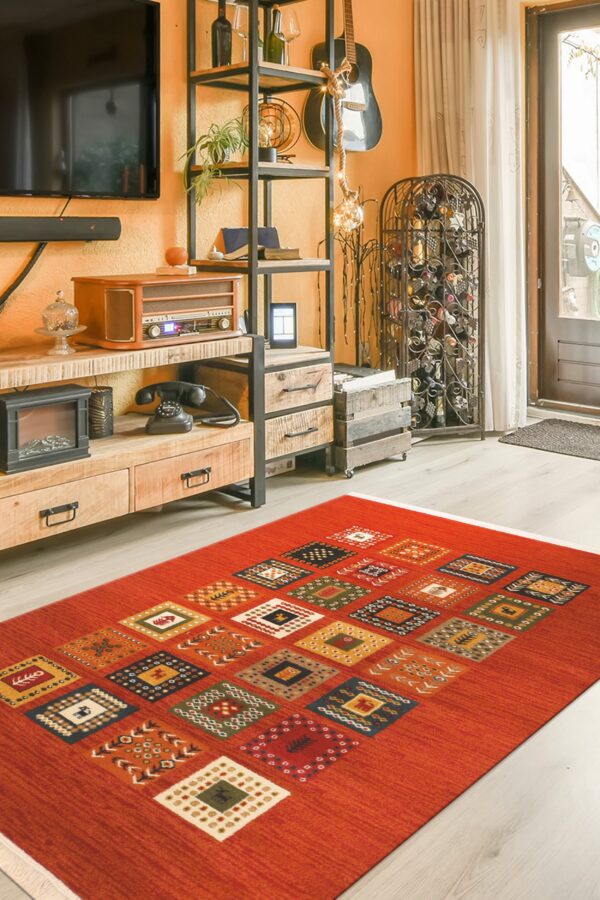 A rust-orange area rug with colorful patterned squares lies on light wood flooring in a cluttered room.