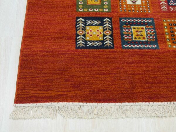Rust rug featuring multicolor geometric squares, cream fringe, and a striated medium pile on light wood flooring.