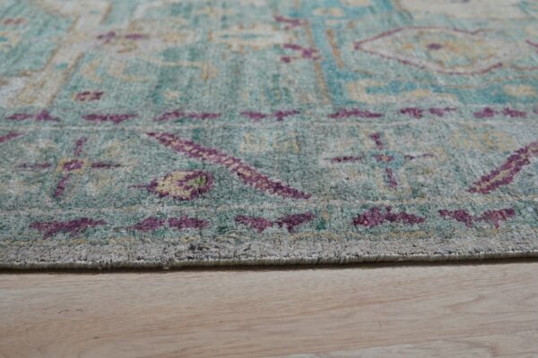Distressed teal and purple patterned rug edge with a medium pile rests on light blonde wood flooring.