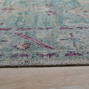 Low pile rug in faded teal, purple, and yellow patterns rests on light blonde wood flooring.