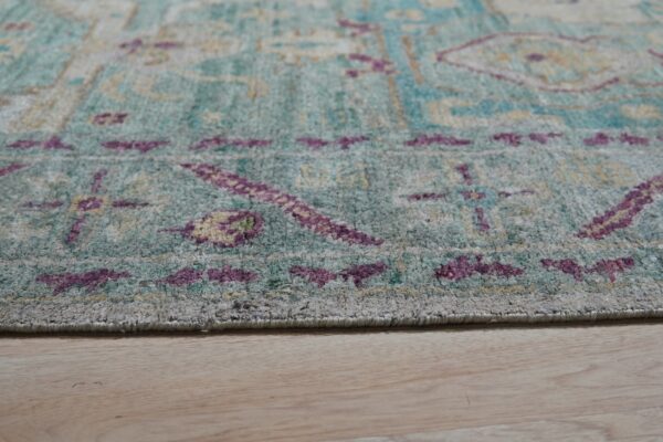 Low pile rug in faded teal, purple, and yellow patterns rests on light blonde wood flooring.