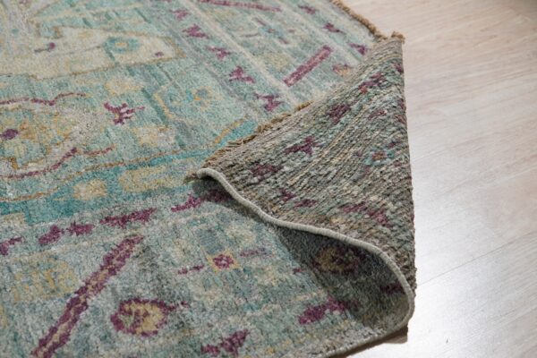 A textured teal, gray, and magenta patterned rug edge is rolled back on light wood flooring.