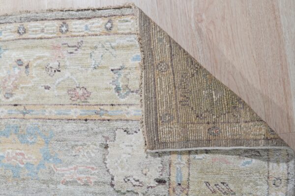 Muted green, cream, blue, and pink patterned rug corner is folded, exposing the woven backing on a wood floor.