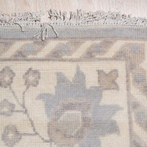 Close-up of a cream rug with a muted gray floral and geometric border and knotted fringe on light wood floors.