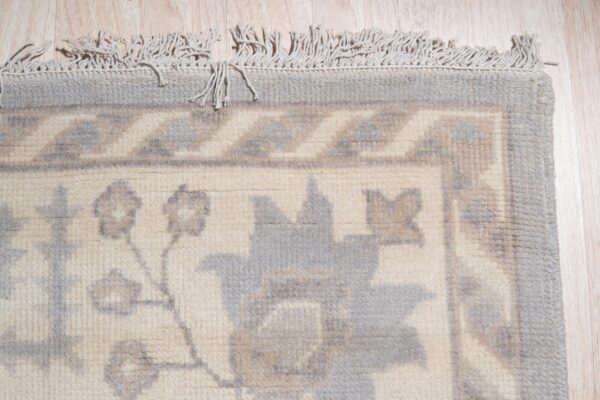 Close-up of a cream rug with a muted gray floral and geometric border and knotted fringe on light wood floors.
