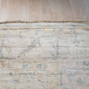 Textured cream rug with fringed edge, showing faded blue and peach patterns on light blonde wood flooring.