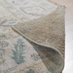The edge of a cream patterned rug with blue and orange details is curled back on light wood flooring.