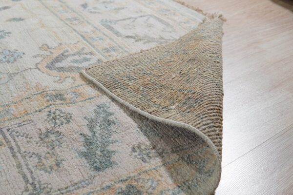 The edge of a cream patterned rug with blue and orange details is curled back on light wood flooring.