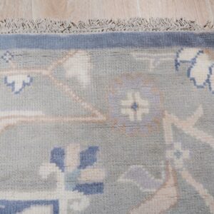Blue-gray traditional rug corner featuring cream and blue floral patterns, a solid border, and fringe on wood flooring.