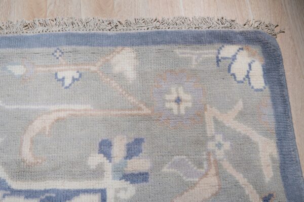 Blue-gray traditional rug corner featuring cream and blue floral patterns, a solid border, and fringe on wood flooring.