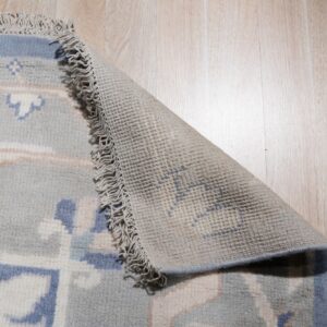 Light blue patterned rug with fringe curled back to show the woven backing on wood planks.