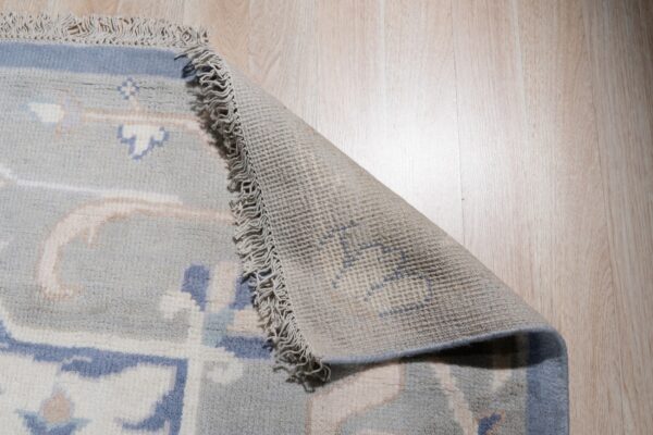 Light blue patterned rug with fringe curled back to show the woven backing on wood planks.