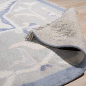 Light blue and beige patterned rug is partially rolled, showing fringe, backing, and light wooden flooring.