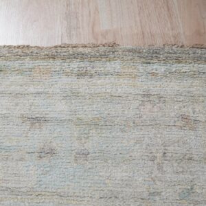 Close-up of a textured, ridged rug in muted cream, gray, and pale blue lying on light wood floor.