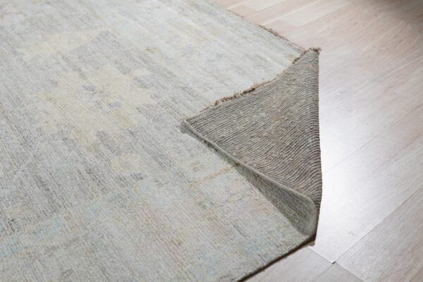 Lightly colored abstract rug lifted at the corner revealing the coarse woven backing on blonde planks.