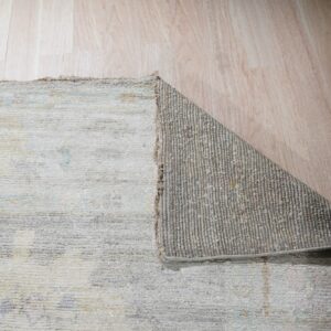 Low-pile rug in pale gray and beige is folded over, exposing the ribbed gray backing on blonde wood.