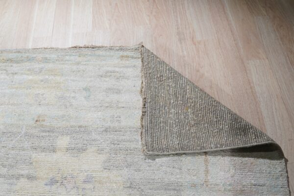 Low-pile rug in pale gray and beige is folded over, exposing the ribbed gray backing on blonde wood.