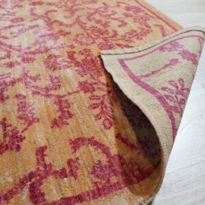 A distressed orange and magenta patterned rug corner is lifted, revealing its woven backing on light flooring.