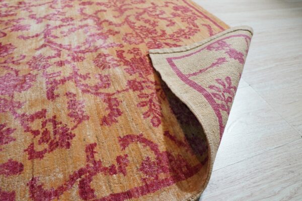 A distressed orange and magenta patterned rug corner is lifted, revealing its woven backing on light flooring.