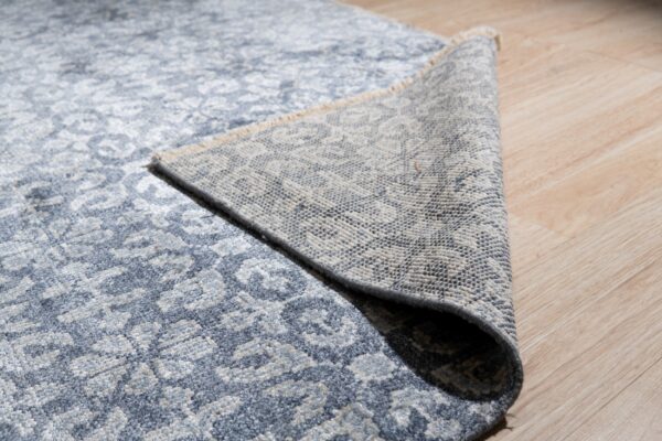 Textured blue and gray patterned rug partially rolled back, showing the woven backing on light wood flooring.