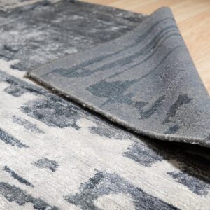 Low pile gray and charcoal rug with abstract pattern, corner lifted on light wood floor.