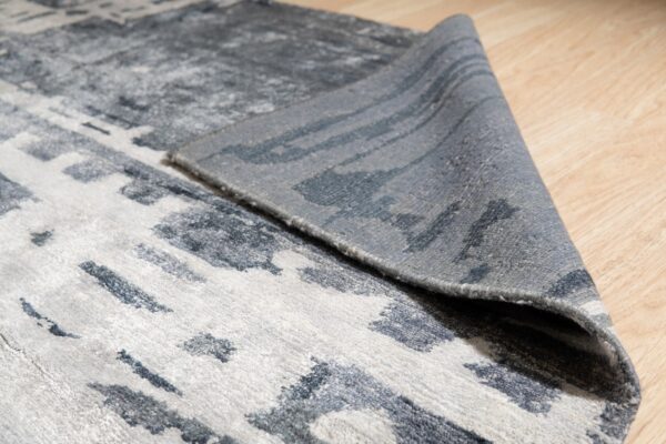 Low pile gray and charcoal rug with abstract pattern, corner lifted on light wood floor.