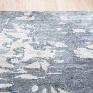 An abstract gray and off-white area rug with contrasting textures rests on light wood planks.