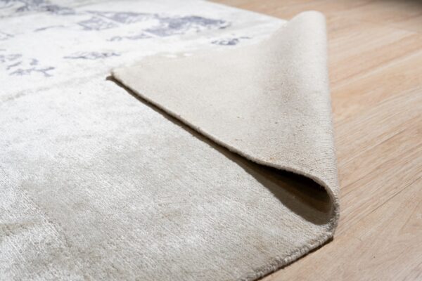 A corner of a cream low-pile patterned rug is folded back on light tan wood flooring.