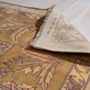 Gold and mauve floral rug with low pile, a corner is lifted revealing the woven backing on light flooring.