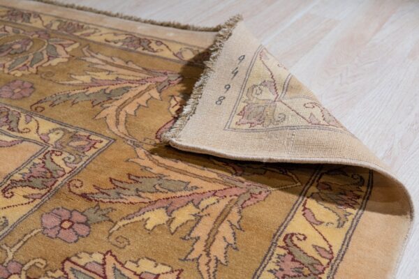Gold and mauve floral rug with low pile, a corner is lifted revealing the woven backing on light flooring.
