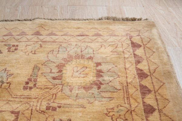 A traditional tan rug with geometric and floral patterns in reddish-brown rests on light wood planks.