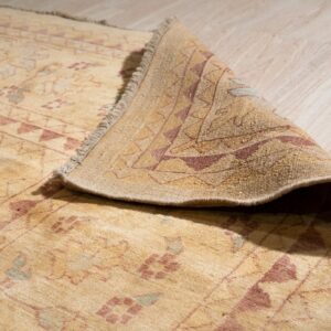 The corner of a patterned beige rug with rust accents is curled, resting on light wooden planks.