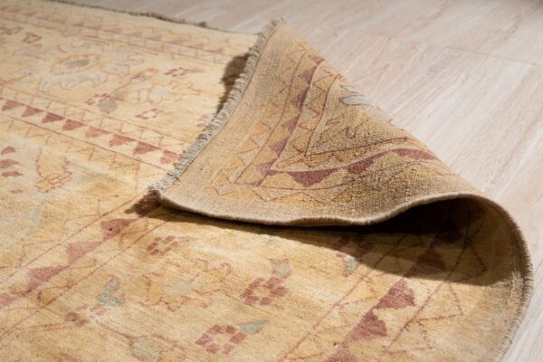 The corner of a patterned beige rug with rust accents is curled, resting on light wooden planks.
