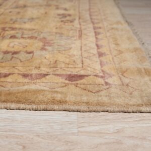 A light golden rug with geometric red patterns and fringe is displayed on pale wood plank flooring.