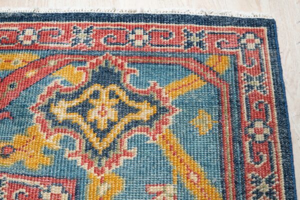 A blue, red, and yellow low-pile rug featuring a dense traditional geometric pattern on light wood floor.