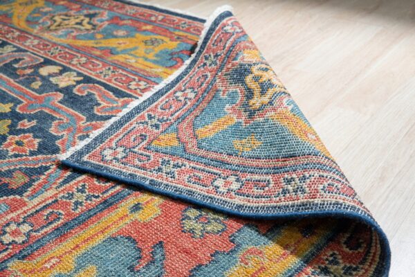 Blue, red, and gold patterned rug folded over light wood flooring, revealing its low-pile tight weave.