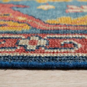 Angled view of a low-pile rug edge featuring geometric patterns in blue, coral, yellow, and beige on wood.