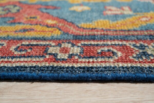 Angled view of a low-pile rug edge featuring geometric patterns in blue, coral, yellow, and beige on wood.