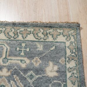 Textured gray-blue rug with cream and teal traditional patterns sits on light wood plank flooring.
